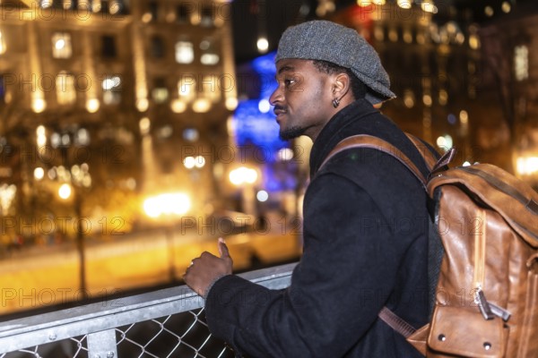 Young black man wearing a cap and coat with a brown backpack, standing on a bridge, thoughtfully watching the illuminated paris cityscape by night with bokeh lights
