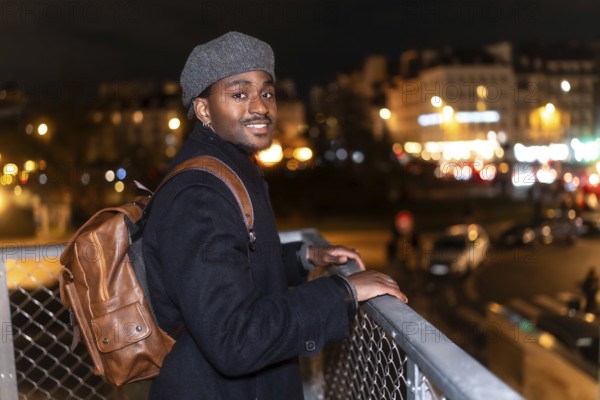 Young black man smiling while looking at the viewer, standing on a bridge overlooking the illuminated city of paris at night, enjoying urban exploration and a relaxed vacation