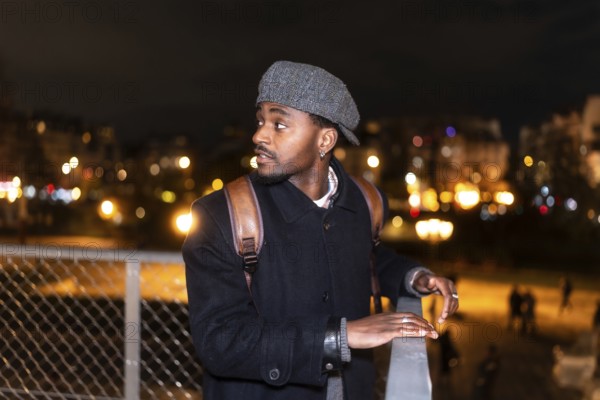 Young man in a flat cap and backpack gazes across a paris bridge at blurred city lights and bokeh nightlife, contemplative traveler soaking in evening urban atmosphere