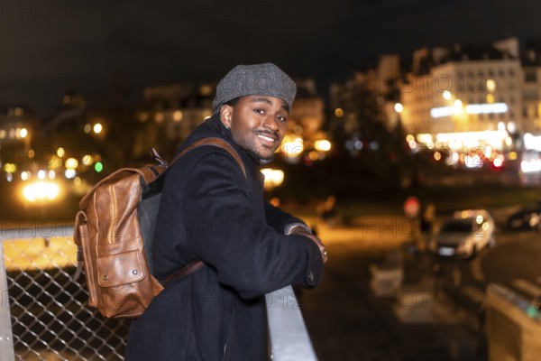 Cheerful male tourist wearing a classic cap and brown backpack, confidently posing on a bridge rail with the vibrant illuminated paris city skyline at night in the background
