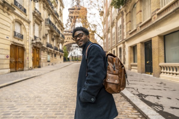 Young african man with glasses and backpack smiles confidently while walking a cobblestone paris street, eiffel tower rising behind historic buildings urban travel joy