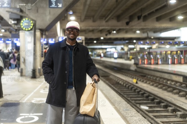 Young black man smiling on a city train platform, wearing a coat and cap, holding luggage and a paper bag while waiting for his train, ready for travel and adventure
