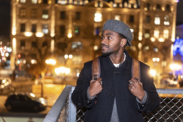 Young man in a beret and coat with backpack pauses on an illuminated paris street at night, gazing at city lights and reflecting on travel, solitude and urban discovery