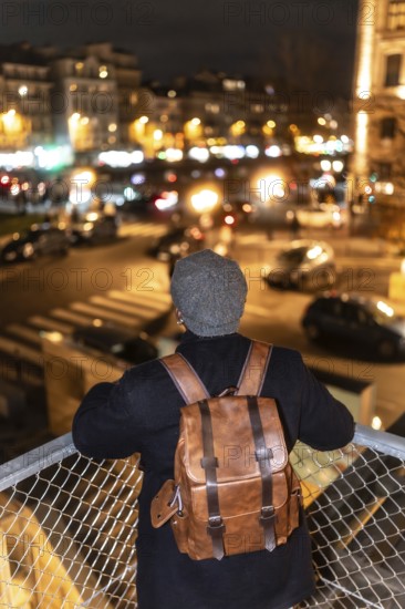 Person with a stylish leather backpack and knit cap viewing the illuminated cityscape of paris, france, from a lookout point, reflecting on urban life and travel