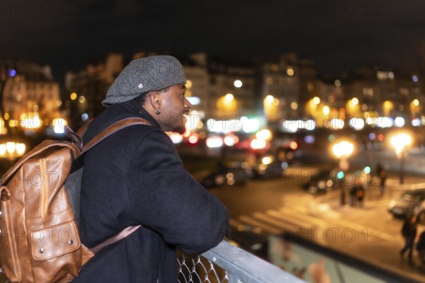 Young black man smiling and enjoying a stunning view of paris street lights at night, wearing a cap and brown backpack while traveling urban cityscape