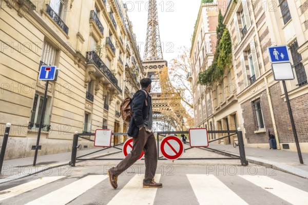 Young man with a backpack walking across a parisian crosswalk, historic buildings and the eiffel tower rising in the sunny autumn background, urban travel and sightseeing scene