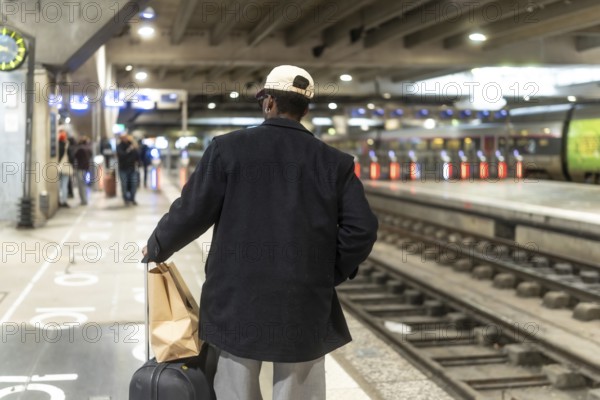 Man standing on a train station platform, seen from behind, pulling a suitcase and carrying a paper bag, waiting for public transport and contemplating his upcoming journey