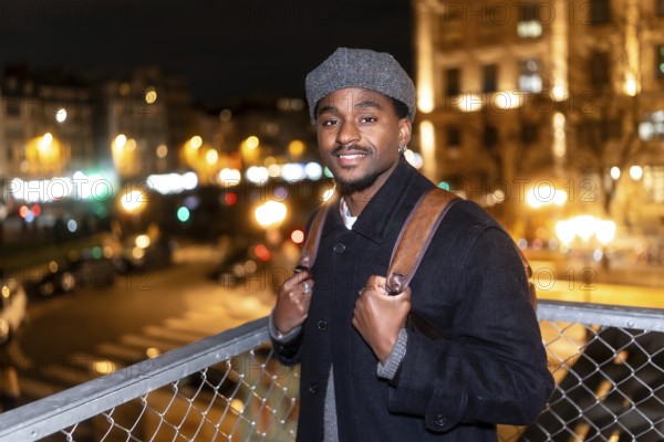 Young african american man smiling, wearing a cap and backpack, standing on a bridge overlooking the illuminated city street lights and buildings in paris during the night