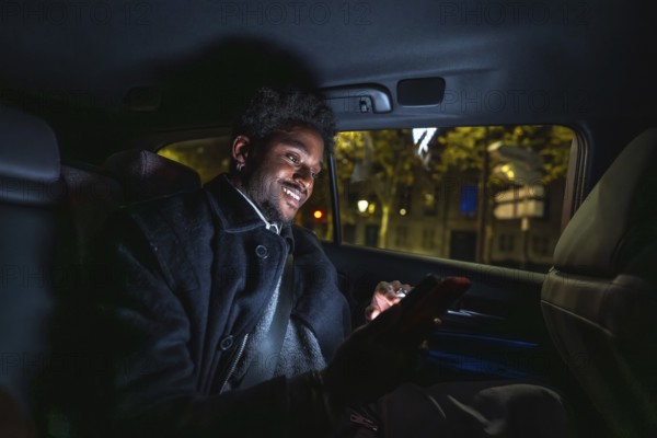 Young black man smiling and connecting with his smartphone, texting or browsing social media applications while traveling in the backseat of a car through the city at night