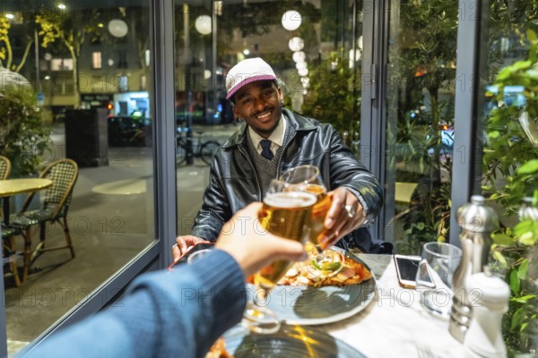 Young man smiling and feeling happy while toasting beer glasses with a friend during an evening dinner out in a modern restaurant setting with city lights reflecting behind them