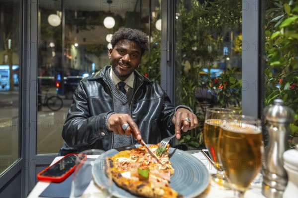 Young man smiling and cutting a delicious pizza slice with a knife and fork, enjoying a relaxing evening meal with a beer in a modern restaurant by the window