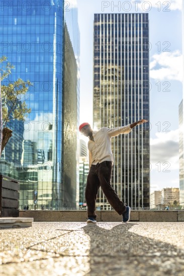 Young man with a red beanie striking a dynamic pose while dancing in an urban cityscape, backlit by sunlight between towering glass skyscrapers on a sunny day