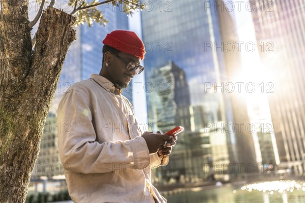Young black man wearing a red beanie and sunglasses checking his mobile phone outdoors, standing by a tree in a modern urban environment with office buildings and sunlight