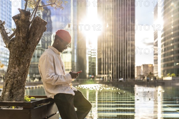 Young black man in casual attire concentrating on his smartphone while leaning by a tree, with modern skyscrapers and shimmering water in the bright evening sun