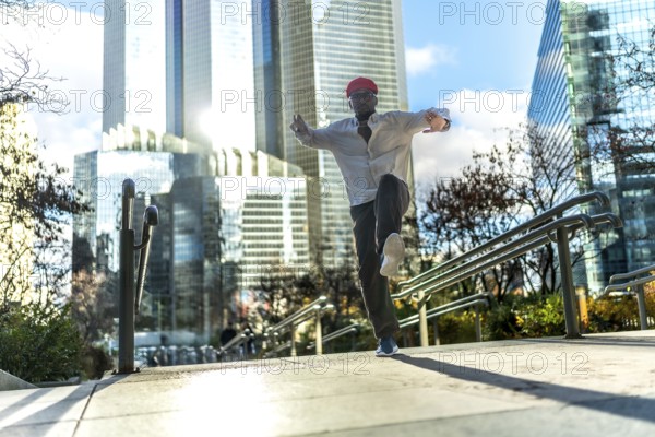 Energetic black man in a red hat and white shirt dancing with joy on an outdoor staircase, expressing freedom and happiness against a backdrop of modern glass skyscrapers and a clear blue sky