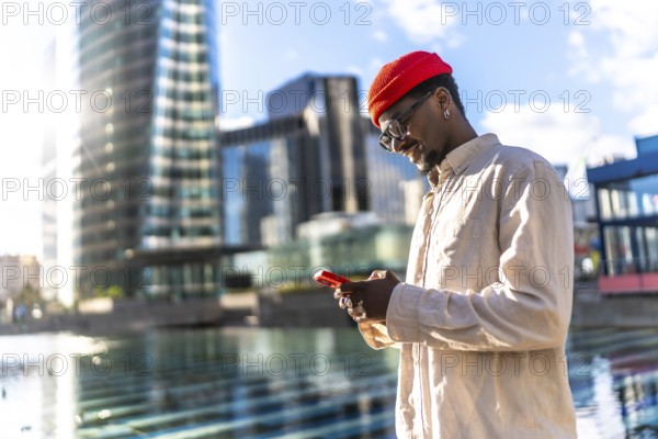 Young black man smiling while interacting with a smartphone outdoors, surrounded by modern city buildings and reflections, representing global communication and urban lifestyle