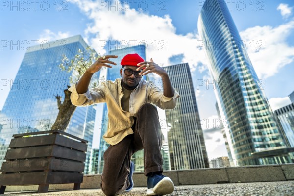 Young man wearing a red beanie and glasses, posing with hands raised in a low angle shot, expressing confidence and modern street style in a city environment with skyscrapers