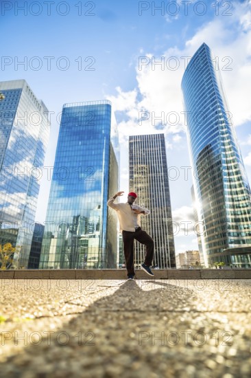 Black man striking a dynamic pose while dancing outdoors in a modern commercial district with glass skyscrapers reflecting the blue sky, expressing freedom and vibrant city life
