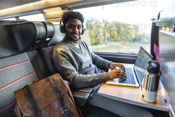 Young african man smiling, wearing headphones and typing on a laptop, working remotely or commuting on a business trip inside a modern train carriage next to a window