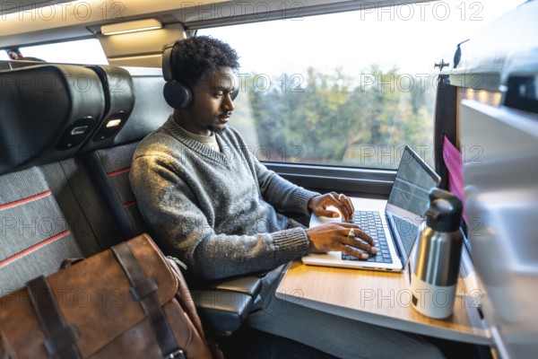 African man traveling on a modern train, wearing headphones and concentrating on his laptop, embodying remote work, digital nomadism, and efficient business travel