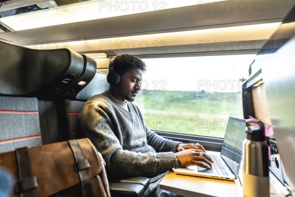 Young man wearing headphones concentrating on his laptop, working remotely or during a business trip while traveling by train, finding focus and productivity in motion