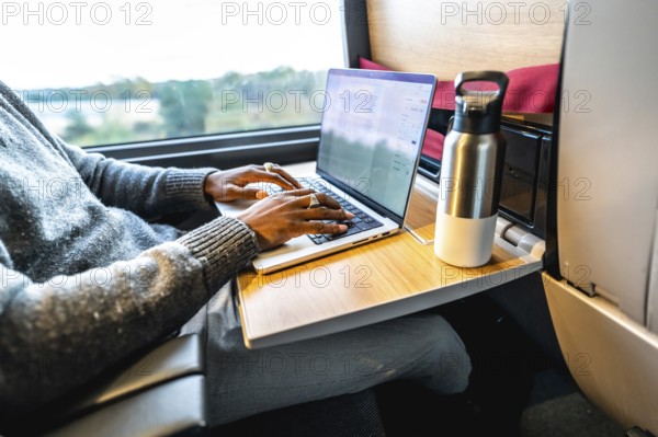 Man typing on a laptop computer while traveling by train, using the fold down tray table as a workspace, combining remote work with a professional journey