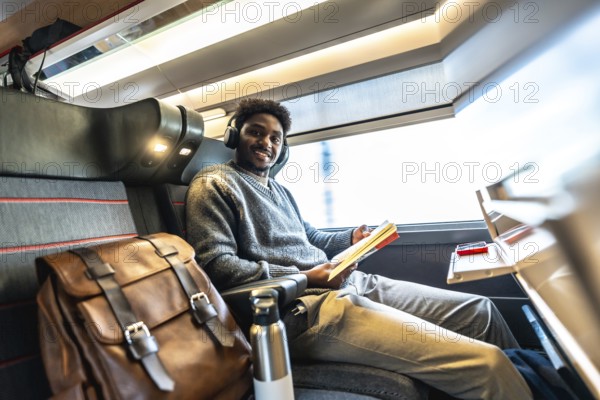 Happy black man wearing headphones and reading a book while traveling by train, sitting comfortably by the window enjoying the journey during a business trip