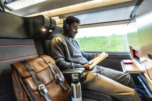Young black man traveling on a modern train, wearing headphones and reading a book, enjoying a comfortable and productive journey with his backpack beside him