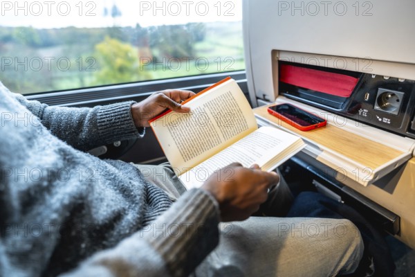 Man reading a book while commuting on a long train journey, using the tray table as a workspace with a phone and charging panel, representing business travel, relaxation, and efficient use of time