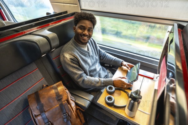Young african american man traveling in a modern train, smiling and looking at the camera while working on his digital tablet, enjoying a comfortable and productive business trip