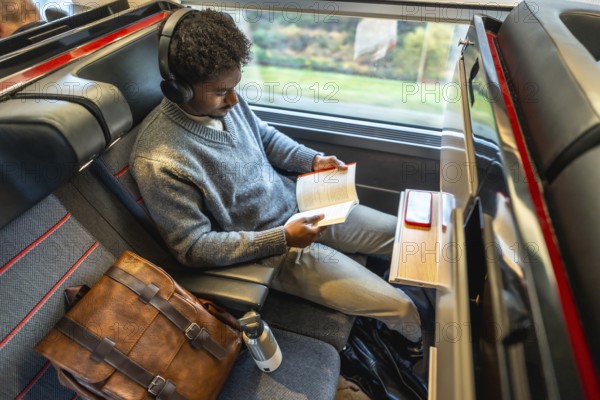 Young professional in headphones sits by a train window reading a book during a high speed intercity business trip, relaxed and focused on personal time and learning