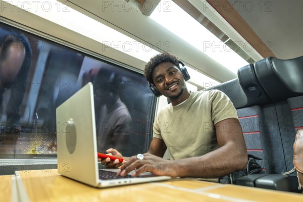 Young black man with headphones smiling while working on a laptop and using a smartphone during a relaxed business trip on a commuter train, digital nomad lifestyle