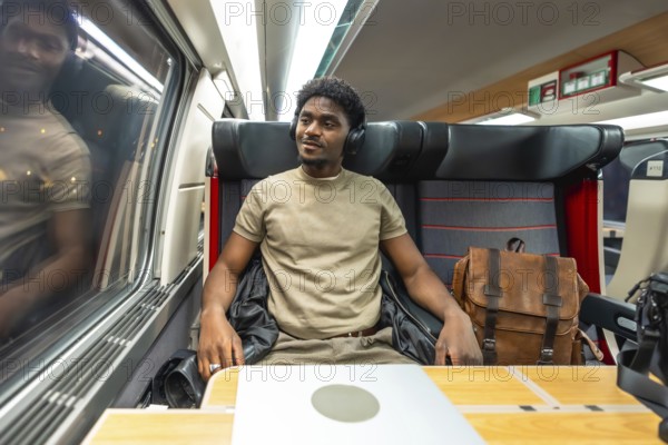 Young black man in headphones and smart casual clothes working on a laptop and gazing out the train window during a calm commute, embodying mobile productivity and travel