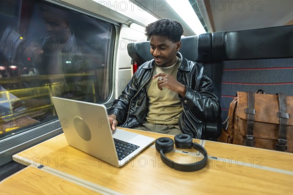 Young man using a laptop and headphones, smiling while traveling by train, reflecting modern business, remote work, and digital nomad lifestyle with connectivity on the go