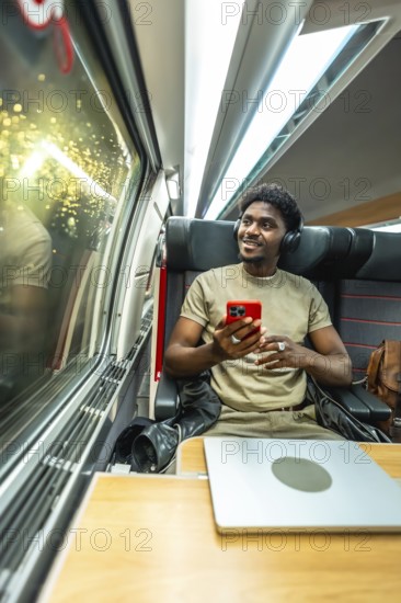 Young man wearing headphones and holding a smartphone, relaxing and enjoying the journey while looking out the window of a modern train, with a laptop on his tray table