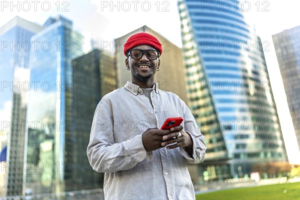 Young black man smiling, looking at the camera while using a smartphone in a modern urban district with glass skyscrapers in the background, enjoying connectivity and tech lifestyle