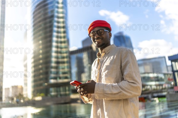 Young man smiling, holding and looking at a mobile phone, standing in a vibrant urban environment with sun drenched modern glass skyscrapers reflecting the sky