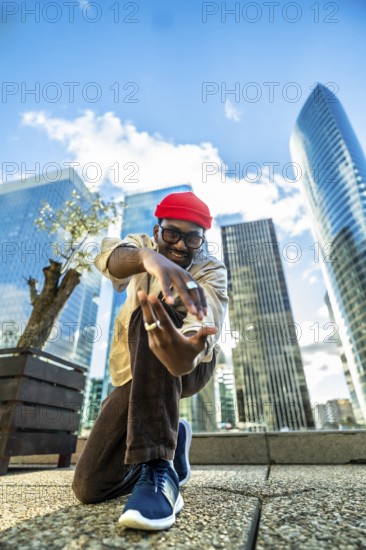Young black man with a red beanie and glasses smiling at the camera, posing and expressing himself with dynamic movements in an urban setting surrounded by modern skyscrapers