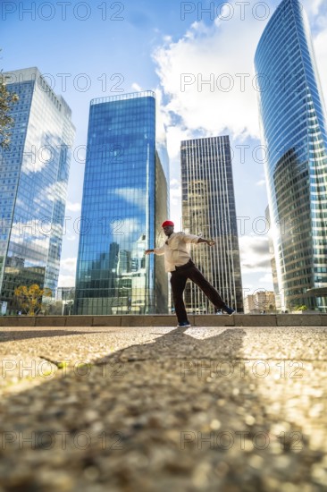 African american man maintaining balance on one leg with outstretched arms, performing street dance in front of modern skyscrapers under a bright sky, embodying freedom and movement