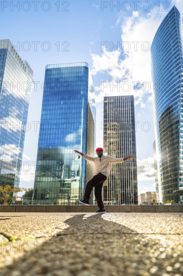 Stylish black man balancing on one leg with outstretched arms, expressing joy and freedom against a backdrop of modern skyscrapers in a sunny urban district