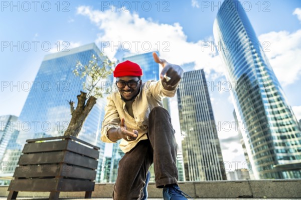 Happy young man wearing a red beanie and glasses, smiling and gesturing towards the camera, standing in a vibrant urban environment with impressive skyscrapers under a bright blue sky