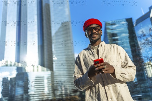 Young man smiling and checking content on his smartphone, linking to technology and modern communication while standing in a business district with glass skyscrapers under a blue sky