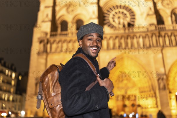 Joyful african american man with a backpack smiling and pointing, experiencing a nighttime tour of notre dame cathedral during his european travel adventure in paris, france