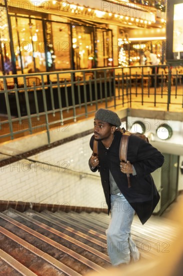 Young black man in beret and coat climbs subway stairs at night, backpack on shoulder, illuminated city architecture and railings blurred behind, urban commuter vibe