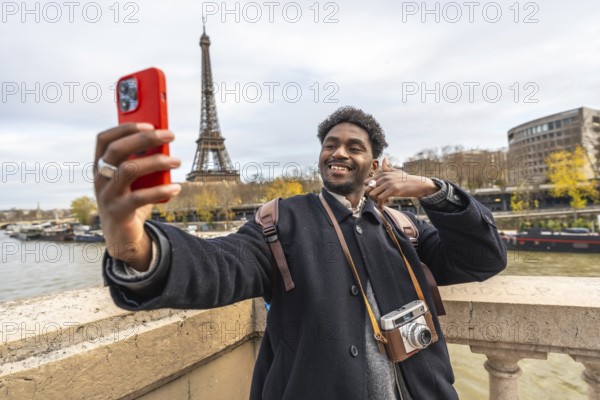 Smiling young man capturing a travel moment with his smartphone, standing on a bridge overlooking the river seine, with the iconic eiffel tower in the background