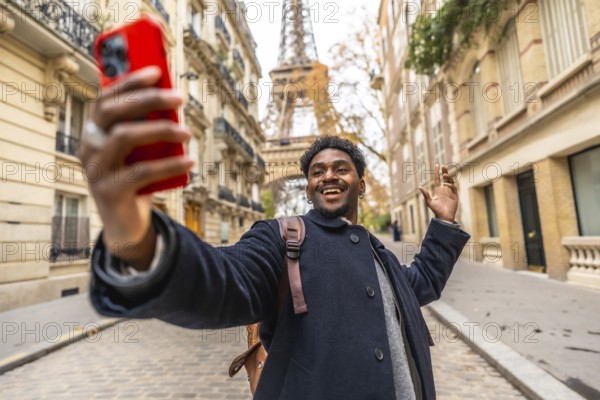 Young black man tourist smiling and recording a video selfie on his phone, waving hand, standing on a parisian street with the iconic eiffel tower in the background while traveling