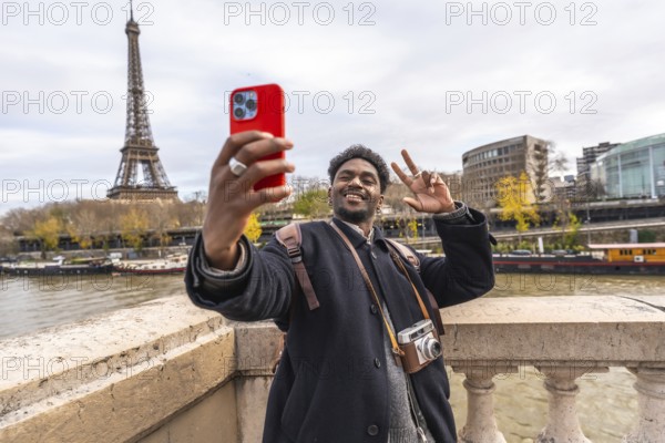 Young man smiling and making a peace sign while taking a selfie with a smartphone, capturing his happy travel experience with the iconic eiffel tower and seine river in the background
