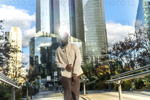 Young man wearing casual street style clothes and sunglasses, looking at camera while posing on steps with modern skyscrapers reflecting bright sunlight in a vibrant urban environment