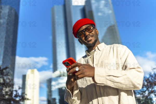 Young black man standing outdoors in a vibrant urban environment, smiling at the camera while holding his smartphone, embodying modern communication and stylish city life