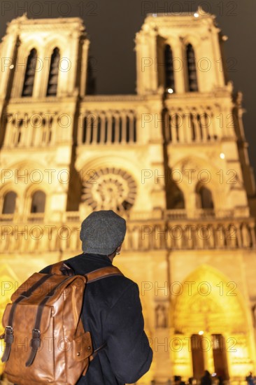 Tourist with backpack and hat seen from behind, gazing at notre dames illuminated gothic facade at night travel, exploration and historic parisian architecture